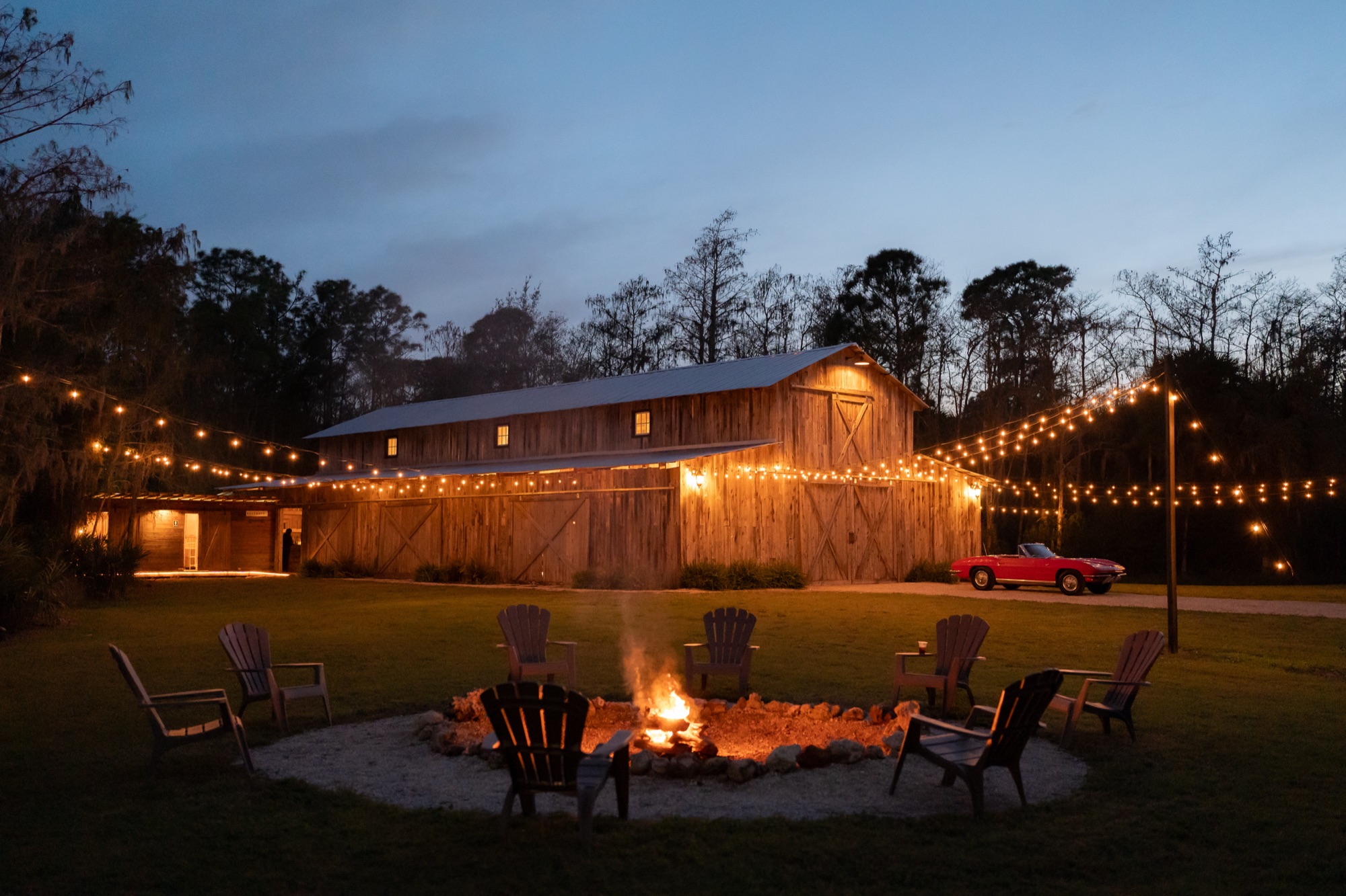 Misty Morning Barn at twilight with string lights, fire pit surrounded by Adirondack chairs, and a classic red Corvette parked beside the barn in Fort Myers, Florida