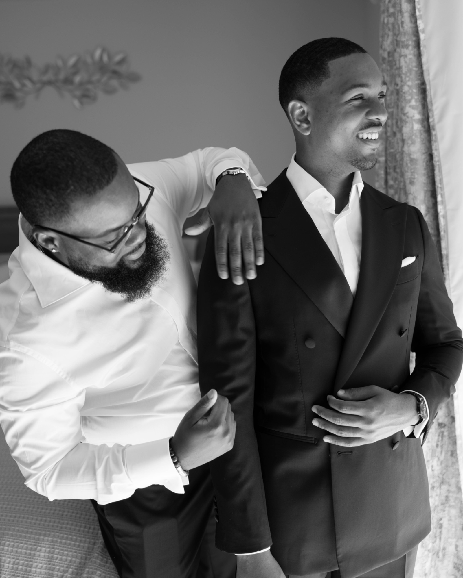 A groomsman laughing with the groom as he helps him dress