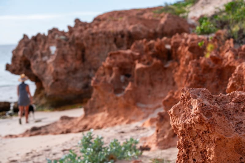 A close-up of weathered red rock formations on a beach, with a blurred woman in a hat and dress walking a dog near the sea in the background.