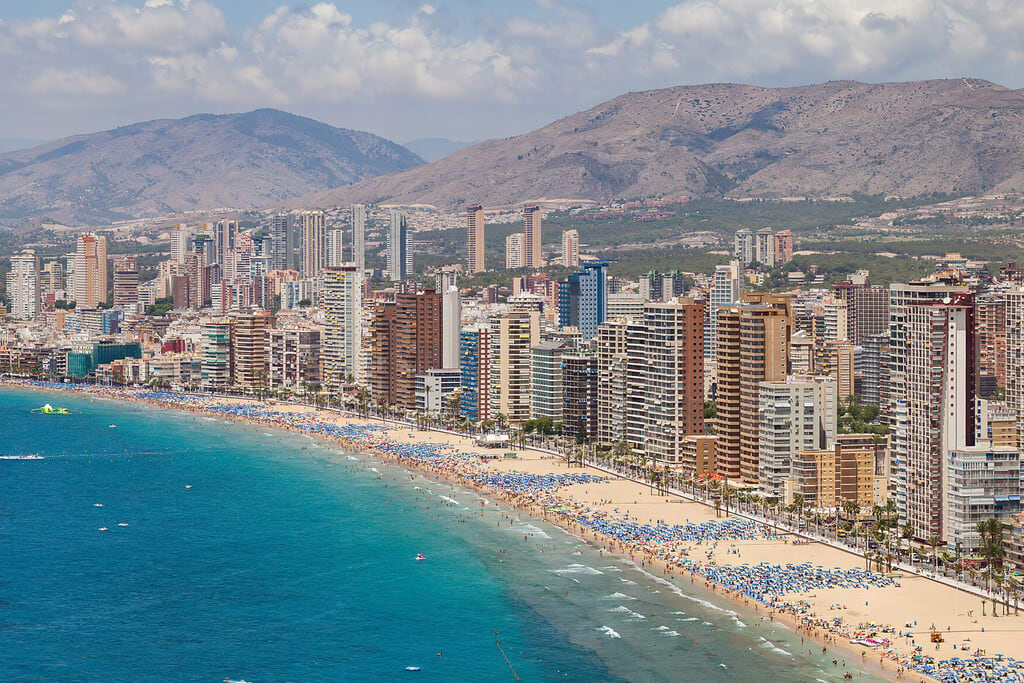 Una vista aérea moderna y limpia de la ciudad de Benidorm con su costa y edificios icónicos, bajo un cielo azul claro.