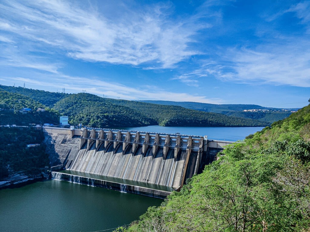 Vista aérea de una masiva presa hidroeléctrica en construcción en un entorno montañoso, con maquinaria pesada y obras visibles.