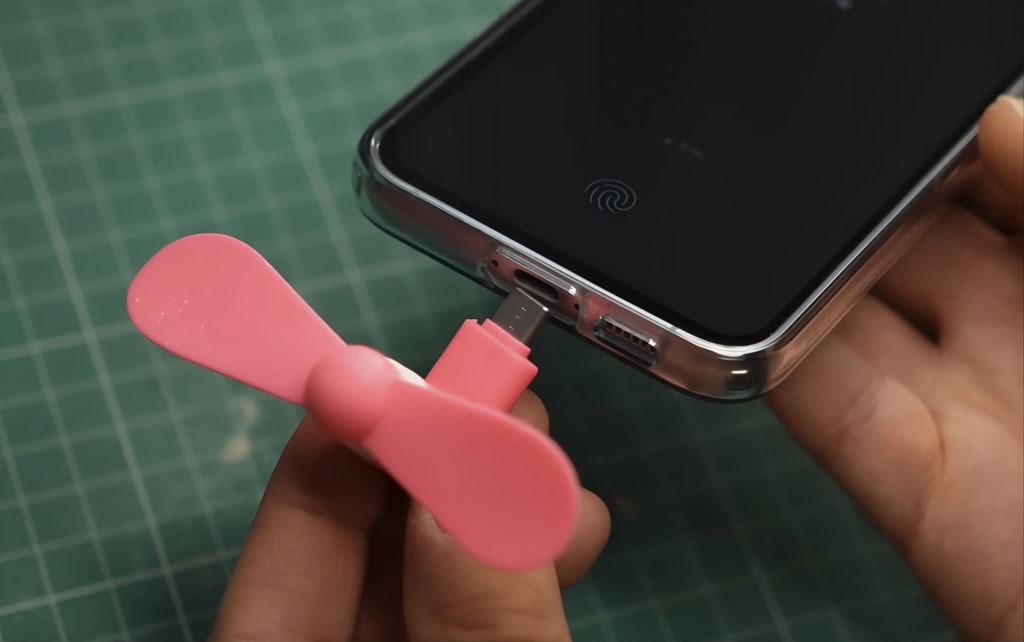 A hand holding a small USB-C fan connected to a smartphone, with a subtle electrical spark graphic indicating danger.