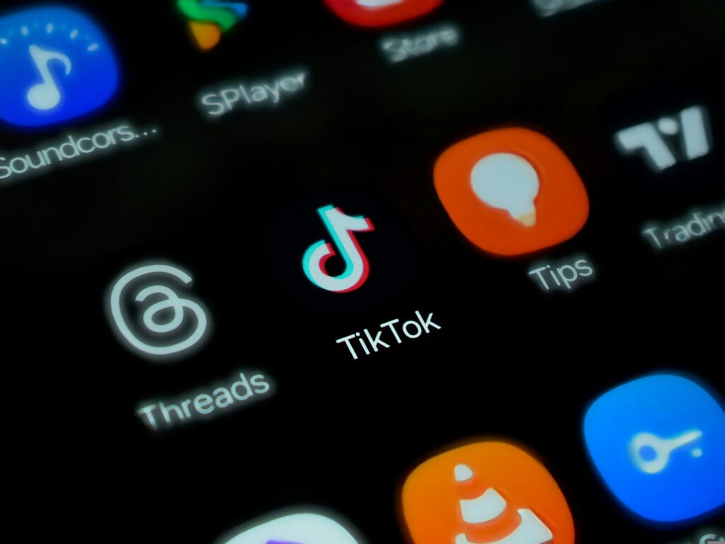 Un primer plano de un teléfono móvil mostrando la interfaz de TikTok, con un fondo desenfocado de luces de neón azules y moradas, sugiriendo la naturaleza digital y a veces oscura de las plataformas online.