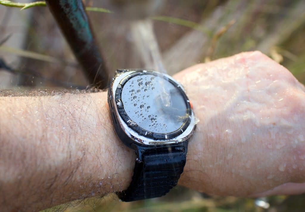 Primer plano del Samsung Galaxy Watch Ultra mostrando su pantalla encendida con la interfaz de Gemini, con un fondo desenfocado de equipamiento deportivo.