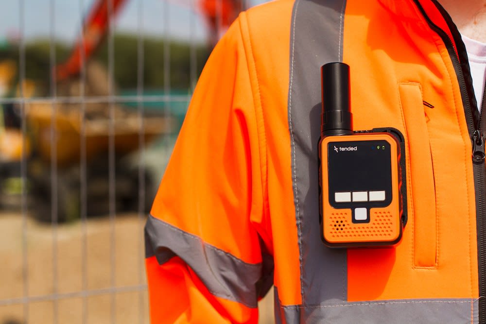 Person in high-visibility orange jacket with a handheld communication device attached to the chest, utilizing geofencing trackside technology. Construction equipment and fencing are visible in the background.