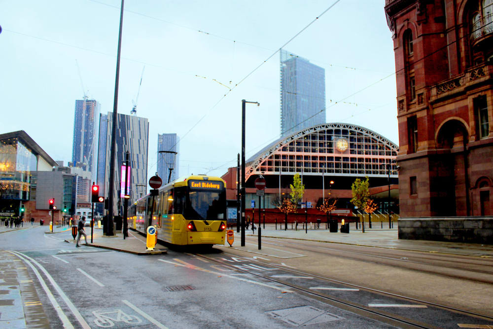 A yellow tram travels through a city street in Northern England, moving forward on levelling up and breaking barriers down. Tall buildings and a large arched structure are visible in the background, while the sky remains overcast.