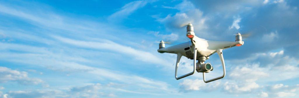 A white quadcopter drone with a camera hovers against a blue sky with scattered clouds.
