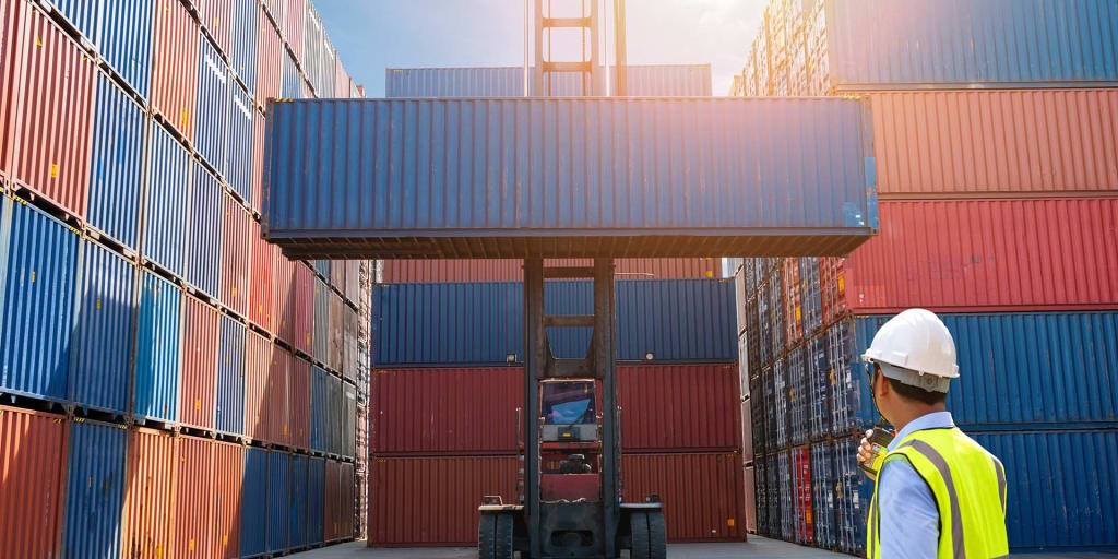 A worker in a hard hat and high-visibility vest oversees a forklift lifting a blue shipping container, surrounded by stacks of colorful containers in a shipping yard.