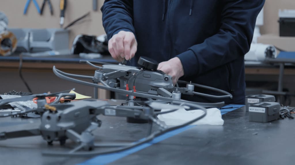 A person in a blue jacket assembles a drone on a cluttered workbench, with another incomplete drone and various tools nearby.