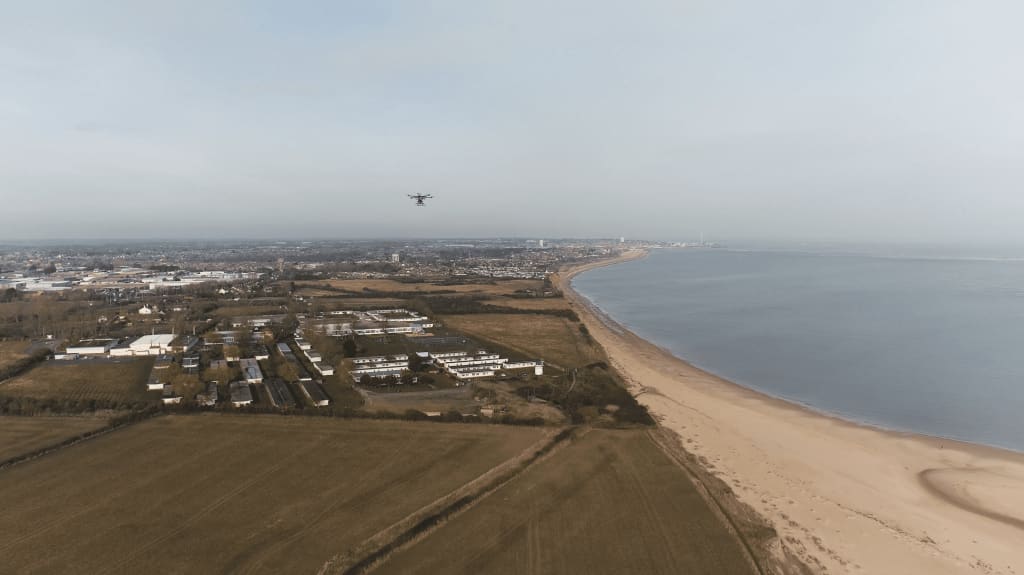 Aerial view of a coastal landscape with a sandy beach, a drone flying overhead, fields, buildings, and a town in the distance.