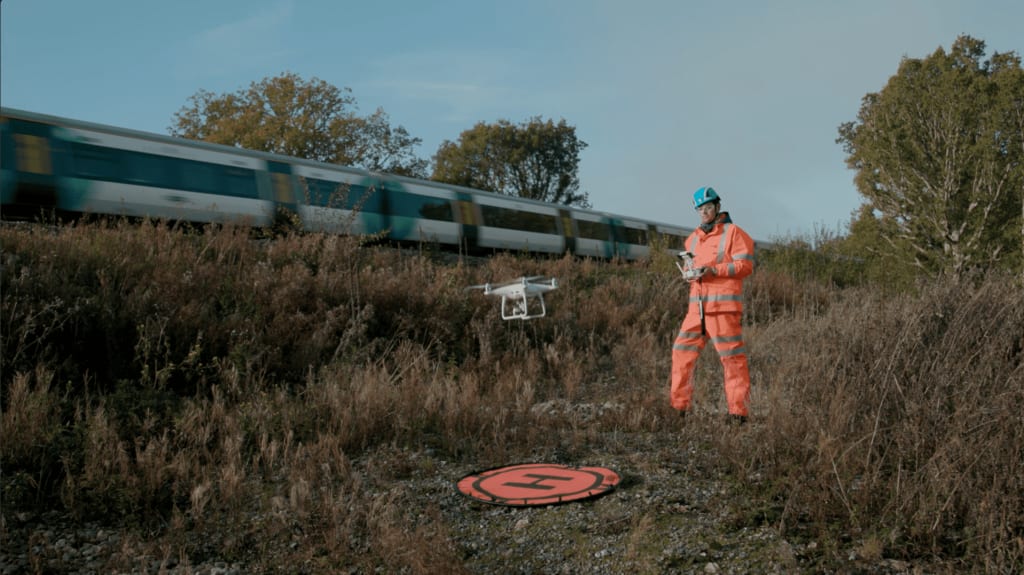 A person in high-visibility clothing operates a drone near a train track, with a moving train in the background. A marked landing pad is on the ground in front.