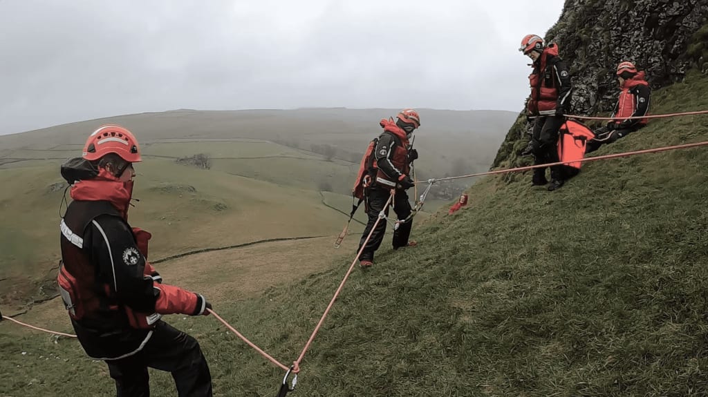 Four rescue workers in helmets and red uniforms conduct a rope rescue operation on a steep grassy hill.