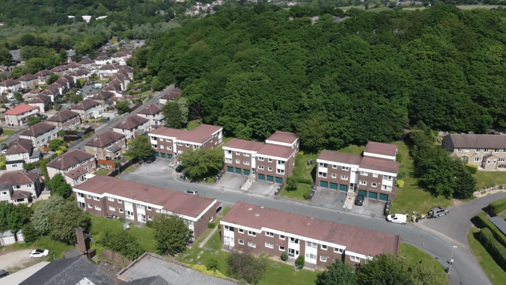 Aerial view of a residential area with multiple two-story apartment buildings, tree-lined streets, and a large wooded area in the background.