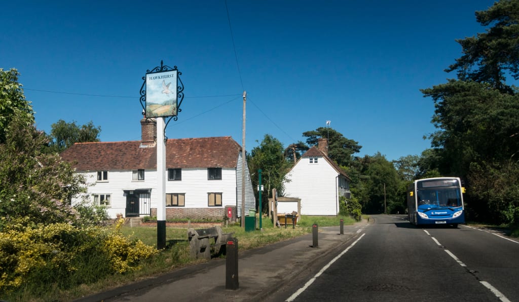 A bus, embodying rural transport, drives down a village road past traditional white houses and a sign for "Smarden" under a clear blue sky.