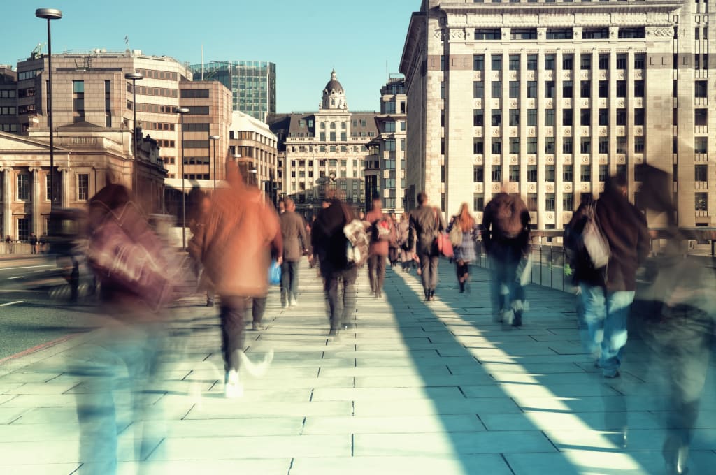 Pedestrians walk across a city bridge, with buildings in the background and a clear sky above. The image has a motion blur effect, giving a sense of movement and busyness.