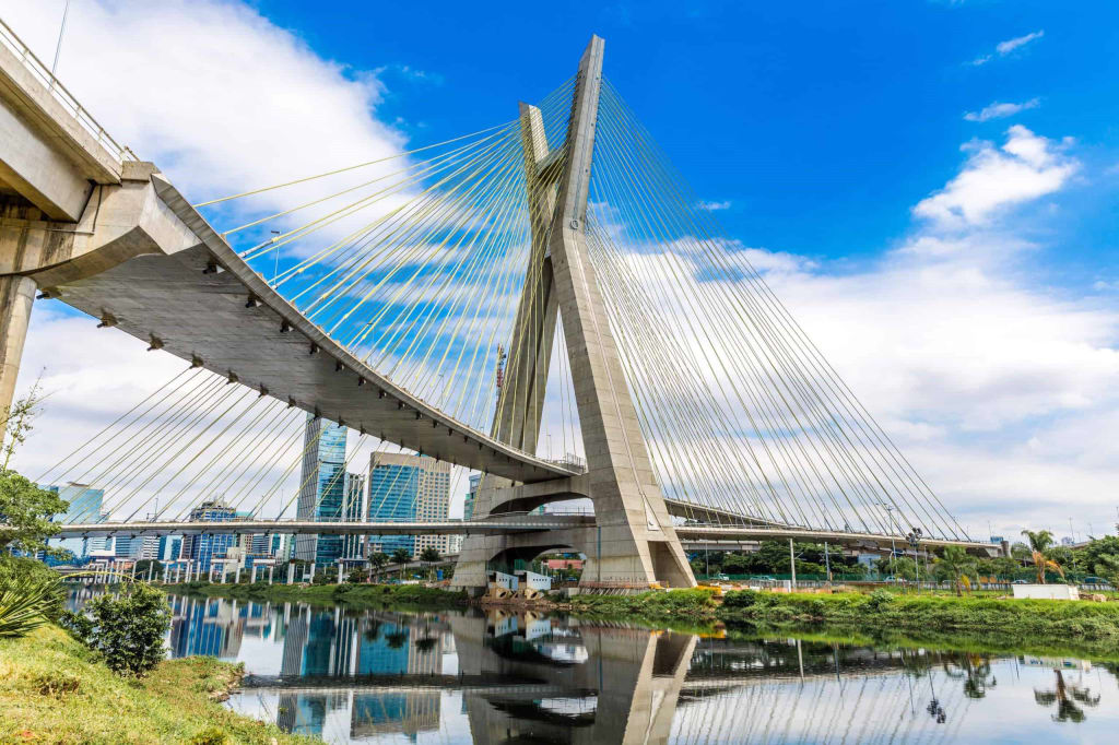 Cable-stayed bridge crossing over a river with skyscrapers in the background against a blue sky with some clouds.