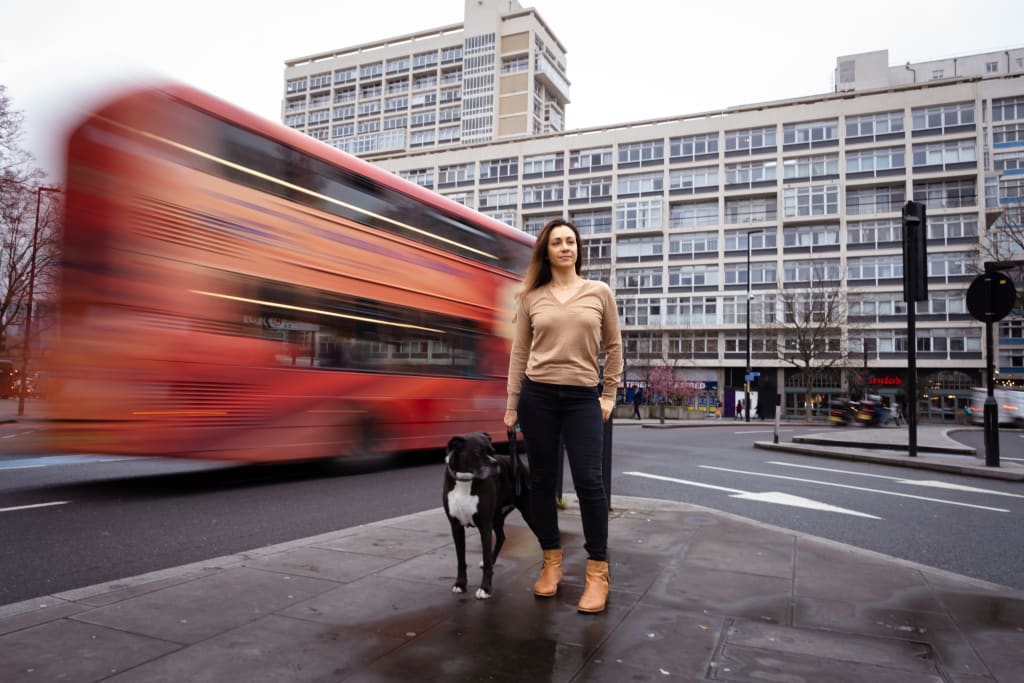 A woman stands on a city sidewalk with her dog as a red double-decker bus, contributing to vehicle pollution, passes by at speed. An urban building is visible in the background.