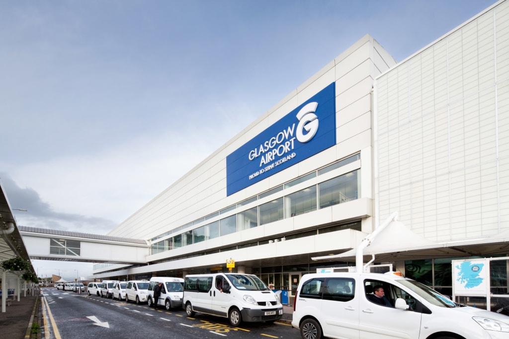 Exterior view of Glasgow Airport terminal building with white taxis lined up in the designated pick-up area, showcasing Glasgow Airport's accessibility features.