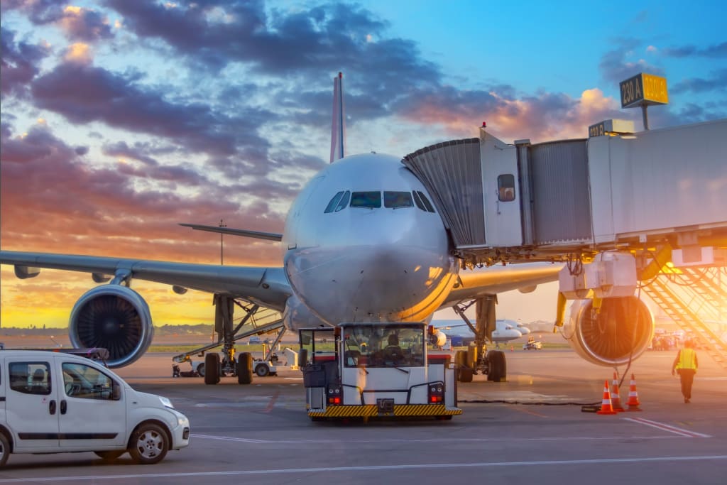 A passenger airplane is parked at an airport gate during sunset at Glasgow Airport. Ground crew and vehicles are servicing the plane while the jet bridge is connected, vividly illustrated in a digital twin representation.