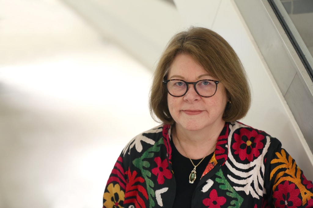 A woman with shoulder-length hair and glasses, wearing a black shirt and a colorful floral-patterned jacket, stands indoors near a staircase, reminiscent of Baroness Brown of Cambridge.