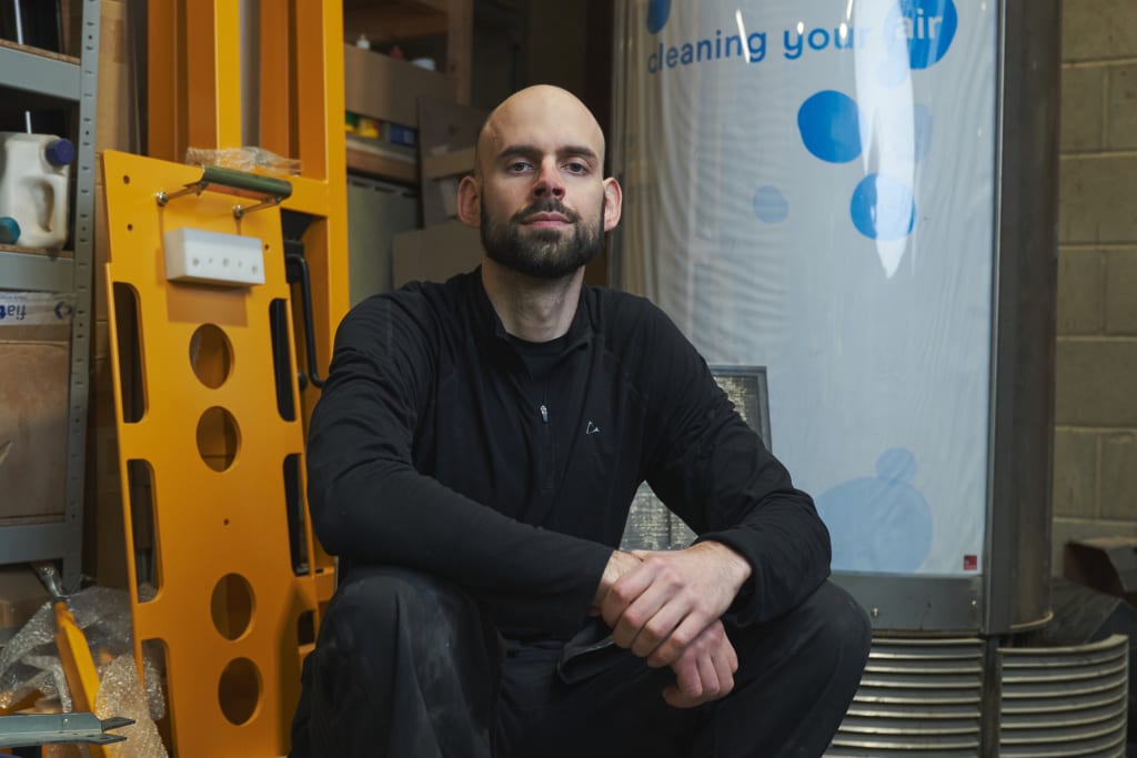 A man with a bald head and a black shirt sits in a workshop surrounded by various tools and equipment, ensuring everything is ready for the day's focus on virtual reality safety.