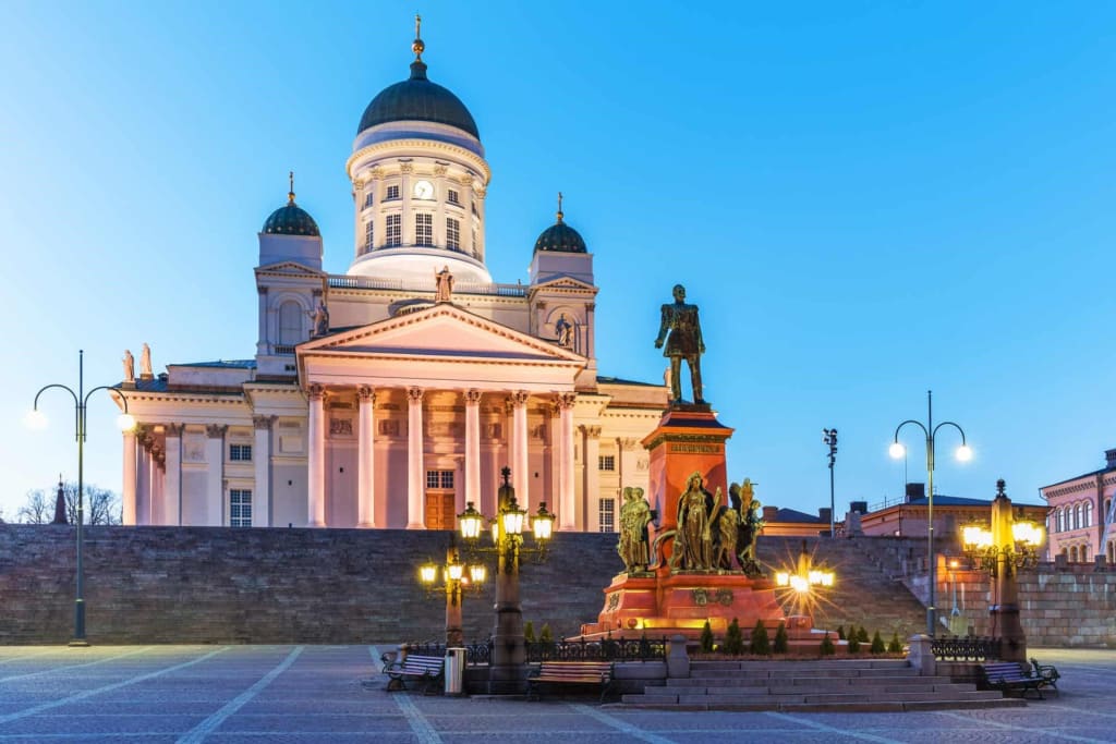 A photograph of Helsinki Cathedral in Finland during dusk, featuring a well-lit statue in the foreground and a clear blue sky.