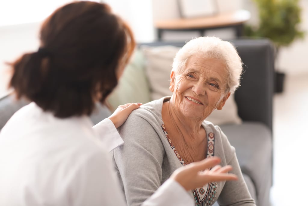 An older woman with gray hair sits on a couch, smiling and gesturing, while a younger person with dark hair in a white coat talks to her, placing a hand on her shoulder.