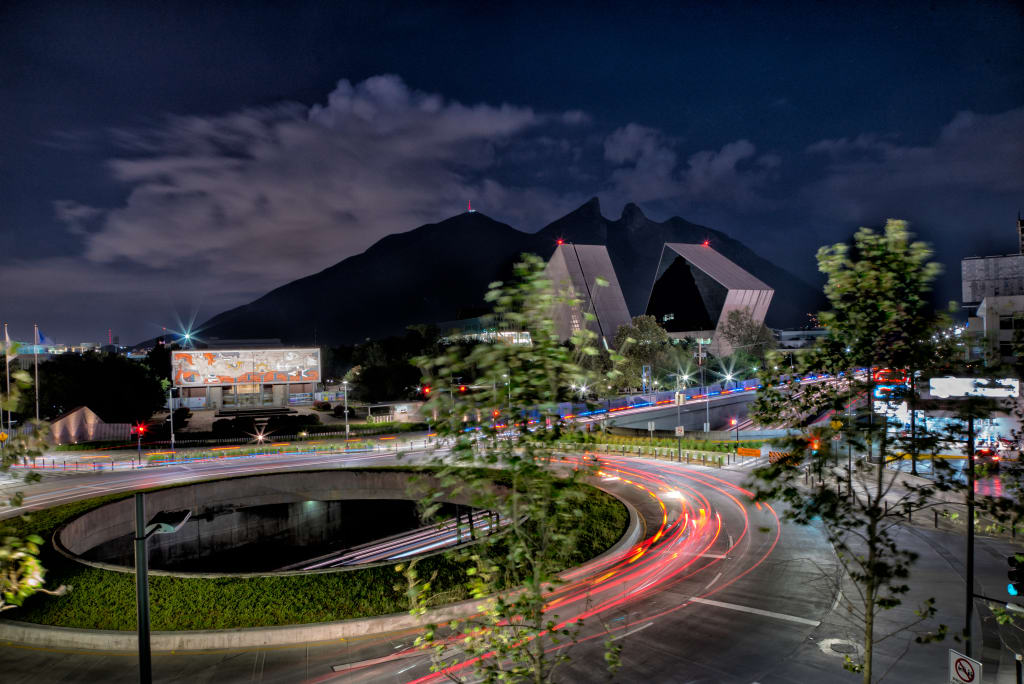 Night view of a modern cityscape with a circular road, blurred car lights, and contemporary buildings. A mountain range is visible in the background under a partly cloudy sky.