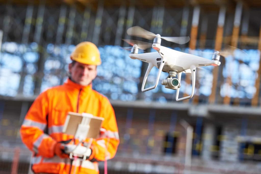 A construction worker in an orange safety jacket and yellow hard hat operates a white drone using a handheld controller at a construction site.