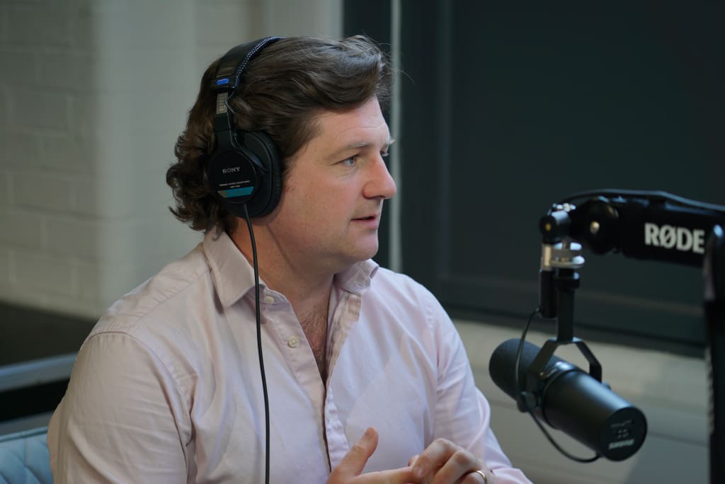 A person with headphones sits at a table, speaking into a microphone. They are wearing a light pink shirt and appear to be in a recording studio, possibly working on creating digital twins for their latest project.