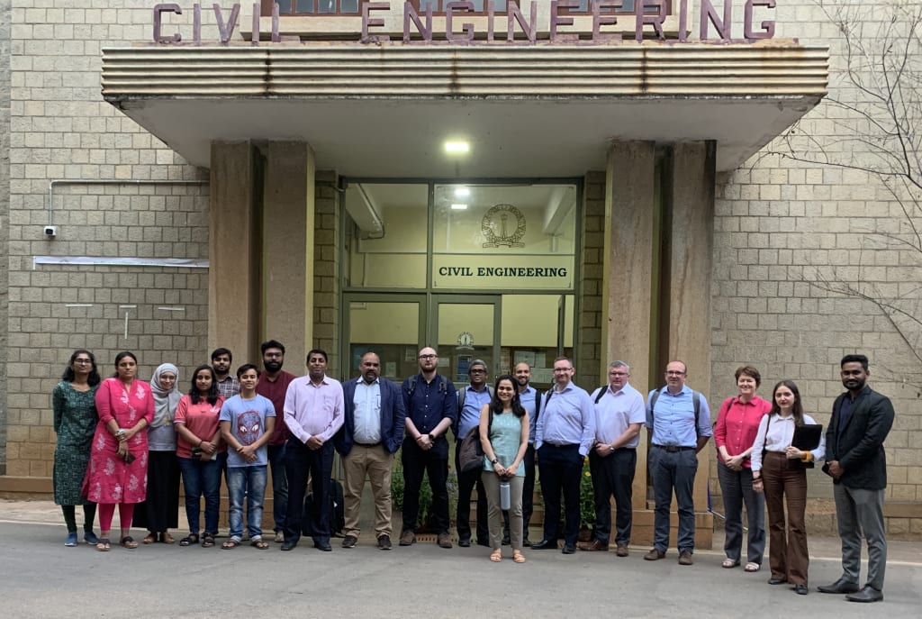 A group of individuals stands in front of a building with a sign that reads "Civil Engineering." The group includes people of diverse ages and attire, posing for a group photo.