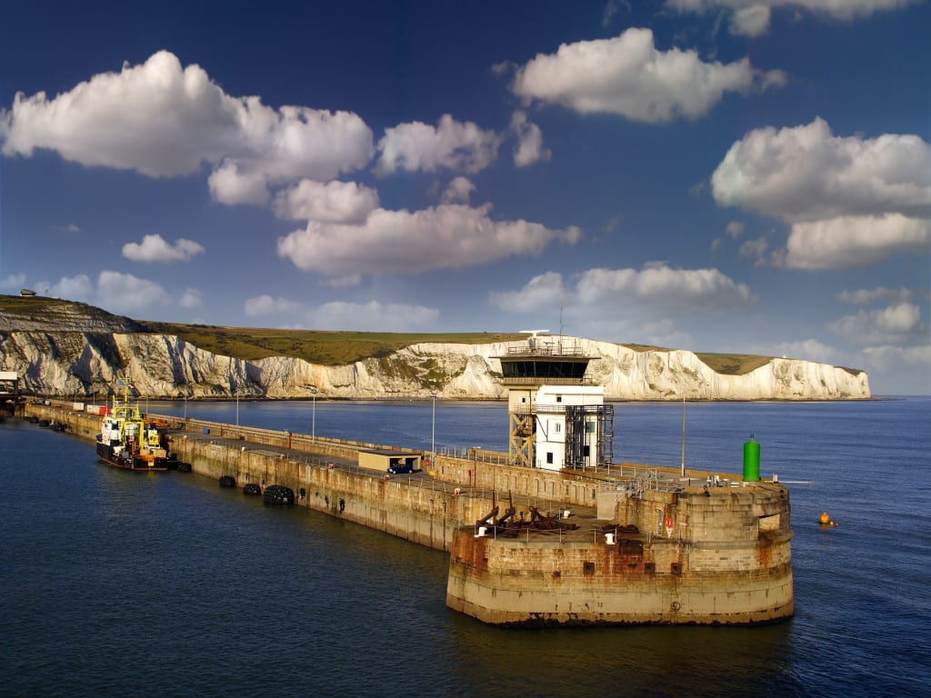 A coastal view featuring a pier and a small docked vessel, with white cliffs and a bright blue sky in the background.