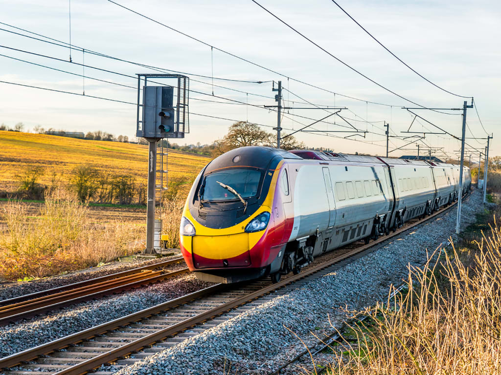 A modern passenger train travels on railway tracks through a countryside landscape with overhead electrical lines.