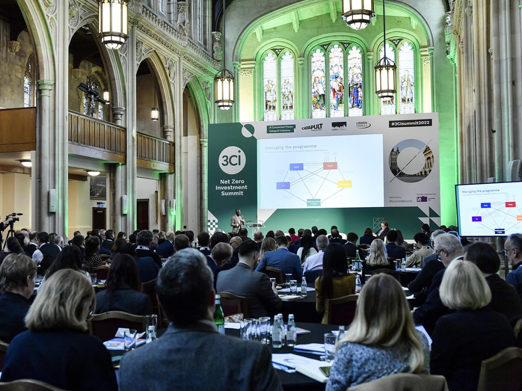 A large group of people attending the Net Zero Investment Summit in a historic hall with stained glass windows. A speaker is presenting from a stage with a screen displaying a diagram.