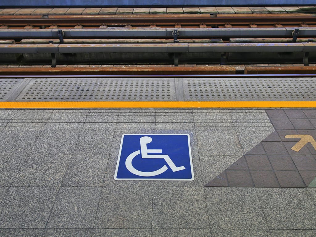 A wheelchair symbol is painted on a train platform near a yellow tactile paving strip, indicating an accessible area for disabled passengers.