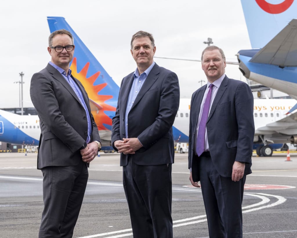 Three men in business attire stand on an airport tarmac with two commercial airplanes in the background.