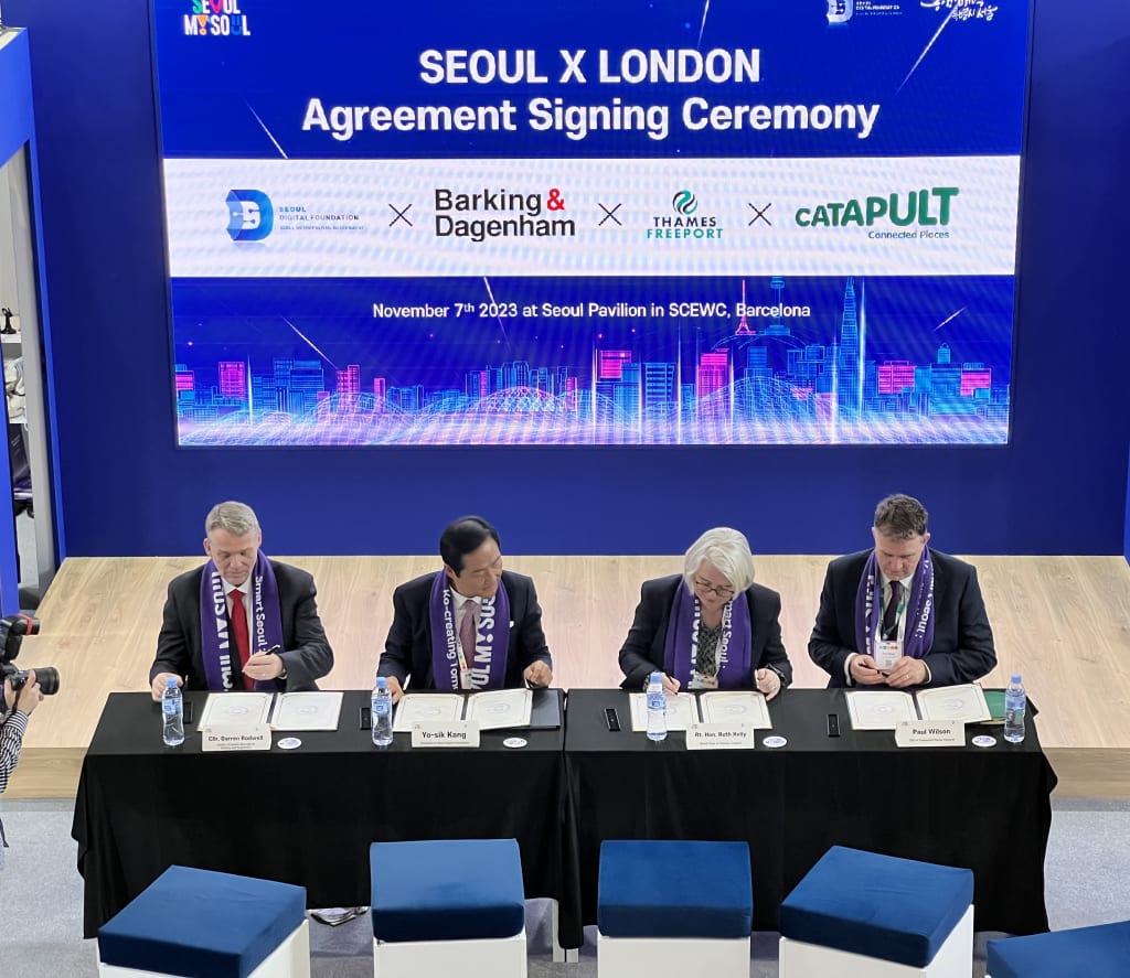 Four individuals are seated at a table signing documents during the Seoul x London agreement signing ceremony, as indicated by the large sign behind them.