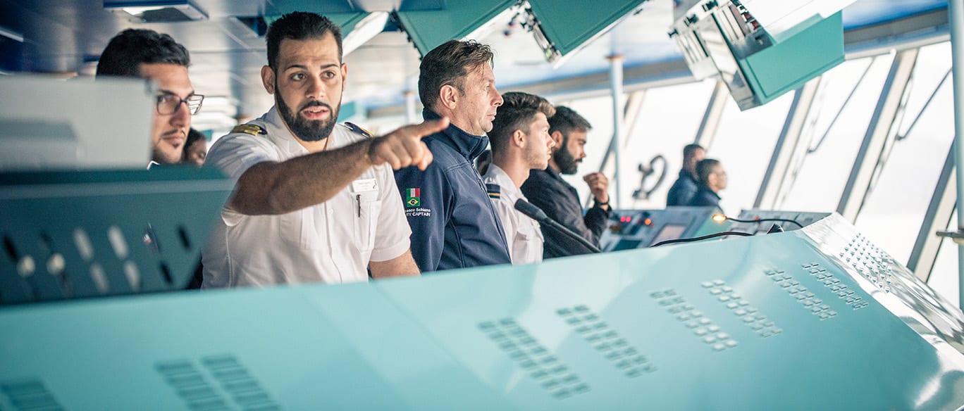 A group of ship officers, dressed in uniforms, stands on the bridge of a ship. One officer is pointing ahead while others observe, surrounded by navigation equipment and monitors.