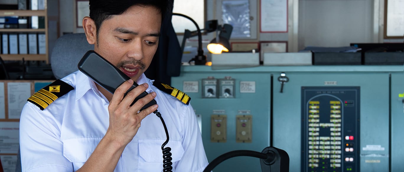 A man in a white uniform with epaulets speaks into a telephone handset in a room with various control panels and switches.