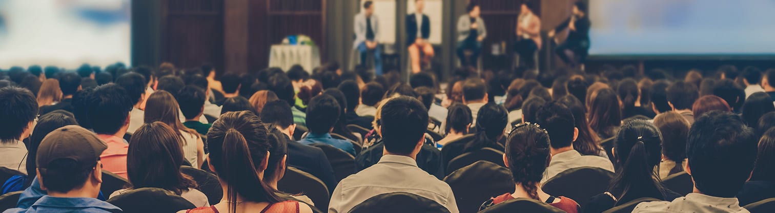 Audience facing a stage with five people seated on a panel, participating in a discussion at a conference or seminar.