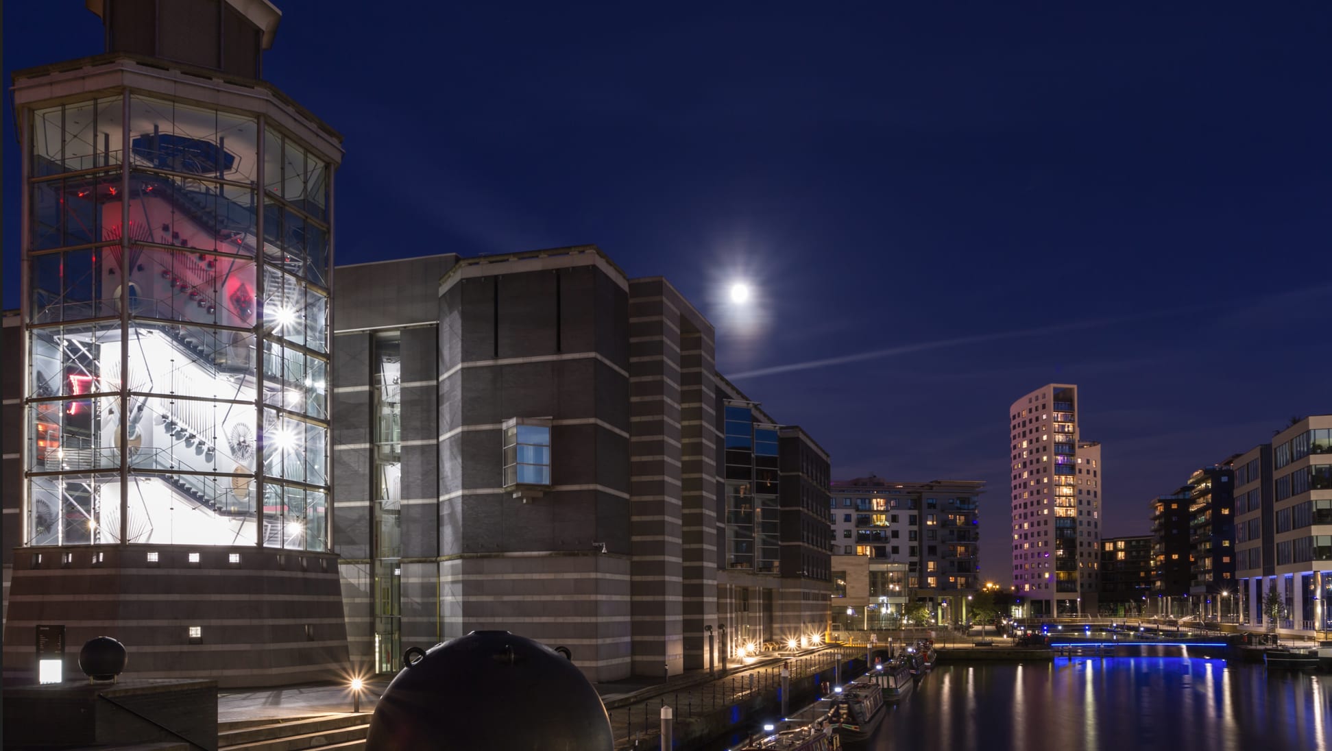 Night view of a modern urban waterfront area, featuring illuminated buildings, a canal, and a full moon in the sky.