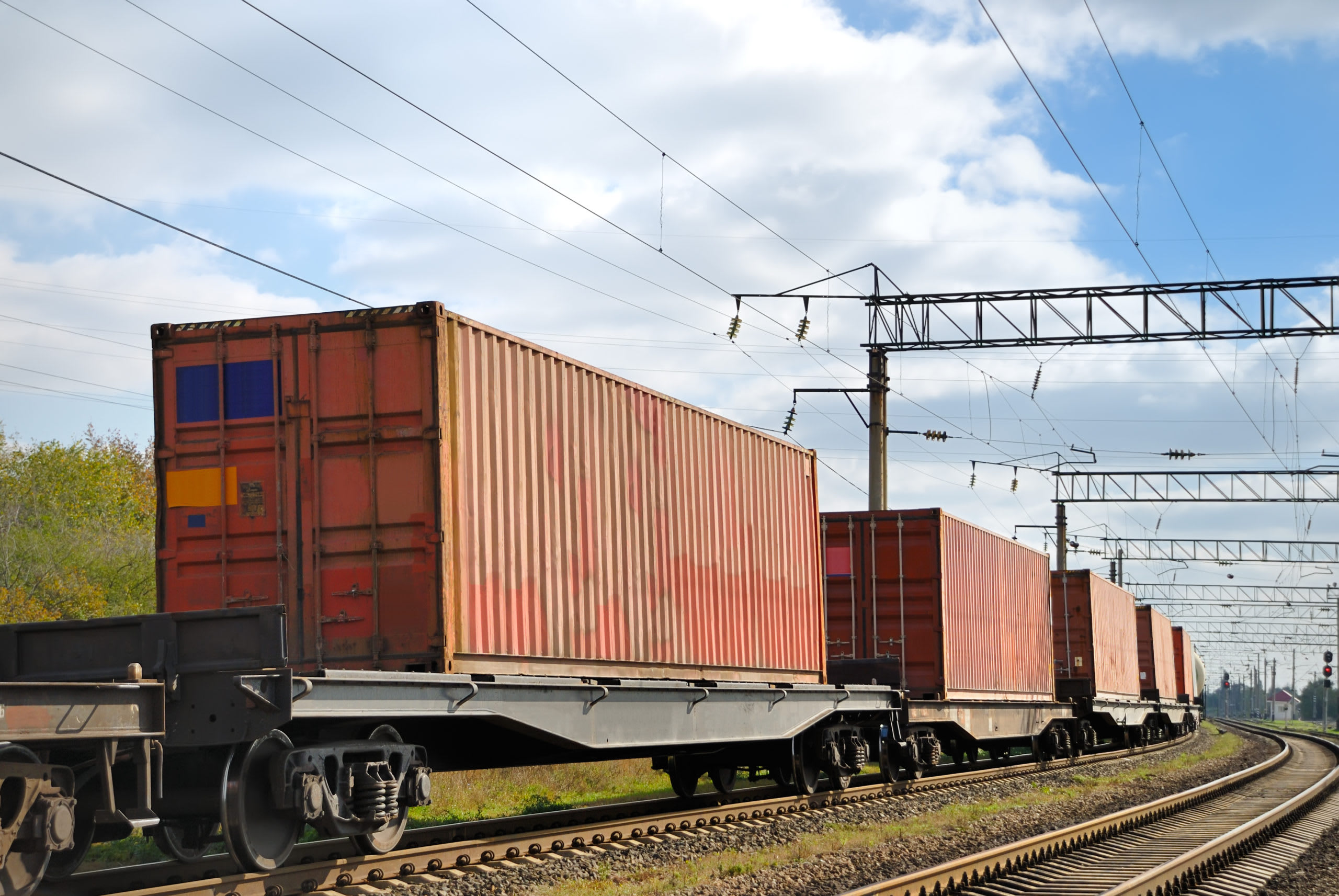 Freight train carrying cargo containers on parallel tracks under a cloudy sky with overhead electrical lines.