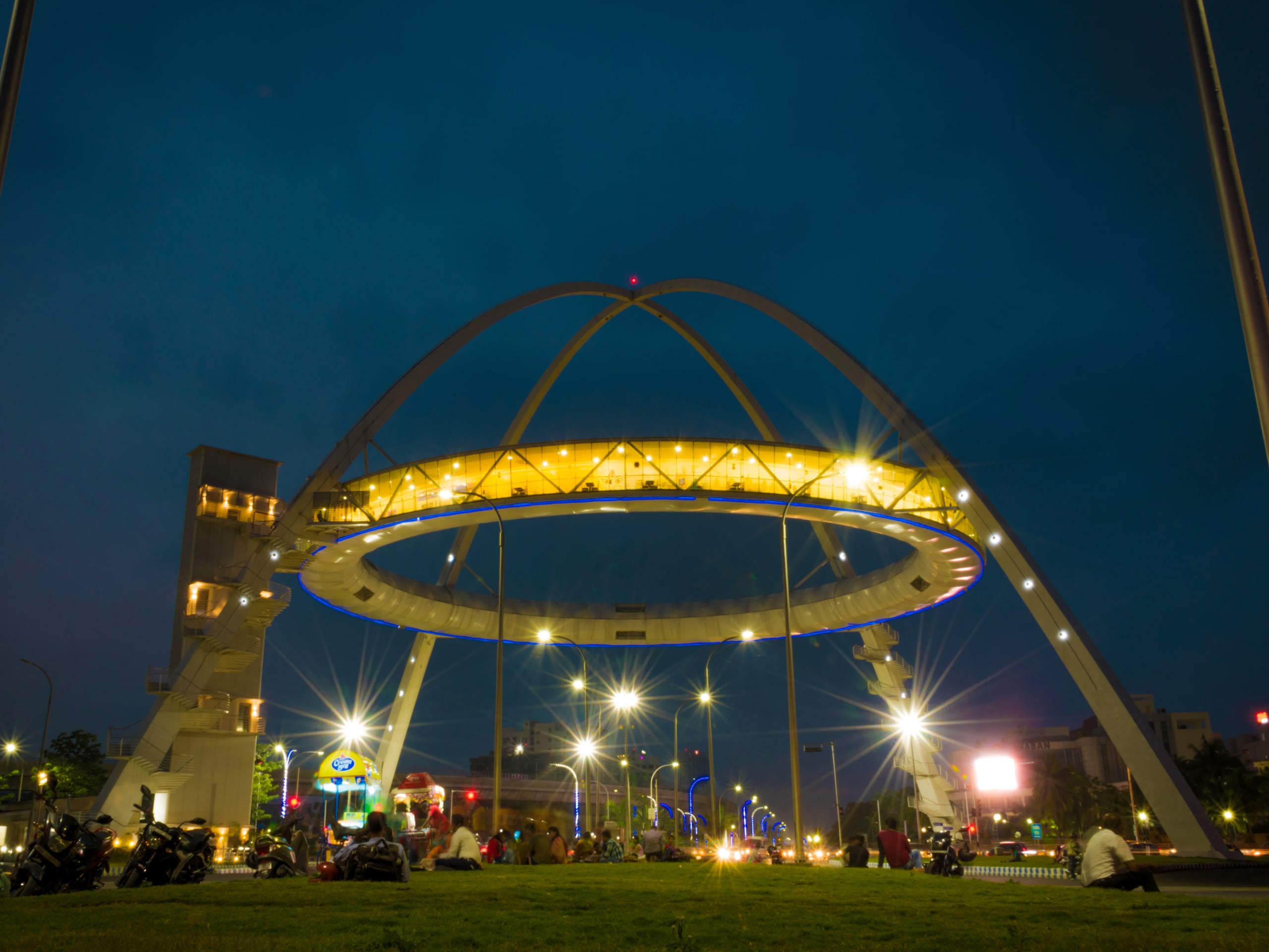 A modern, illuminated arch structure with streetlights glows under the nighttime sky. Buildings stand tall in the background while people and vehicles, including micro-mobility options, navigate near the structure.