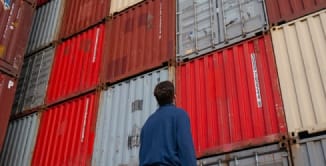 A person in a blue jacket stands in front of a stack of colorful shipping containers, looking up at them.