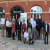 A group of people standing in front of a red brick building with a "Welcome" sign and a banner, posing for a photo. Some are standing, while a few are crouching or seated in front, showcasing the camaraderie at the DIATOMIC Accelerator event.