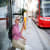 An elderly man with sunglasses and a hat takes a photo with his phone while sitting at a tram stop. A red tram is approaching in the background.