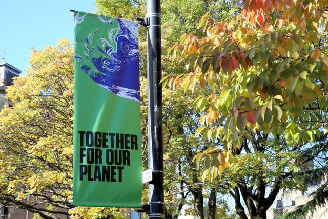 A green banner with an image of Earth and the text "TOGETHER FOR OUR PLANET" hangs on a pole, surrounded by trees with green and yellow leaves.