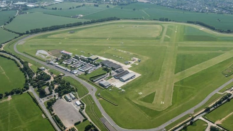 Aerial view of an airfield with runways, green fields, several buildings, and roads connecting various facilities on the premises. Surrounding areas consist of more green fields and a small residential area.