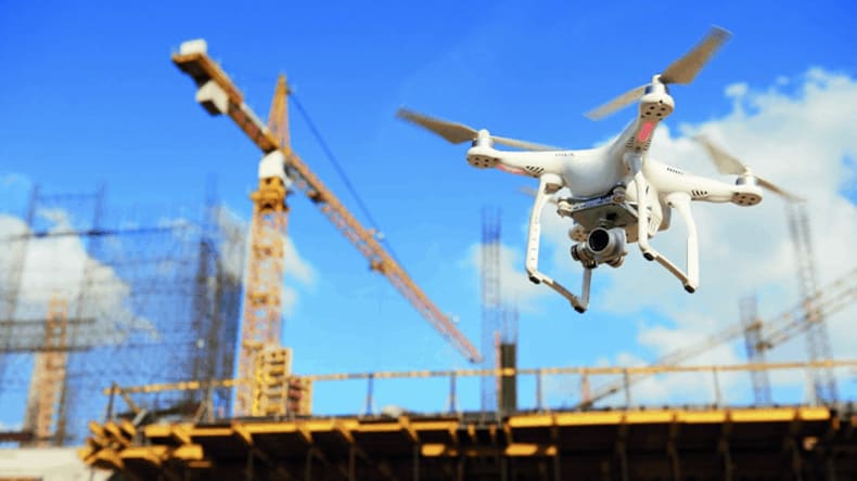 A white drone flies near a construction site with cranes and partially built structures visible against a blue sky.