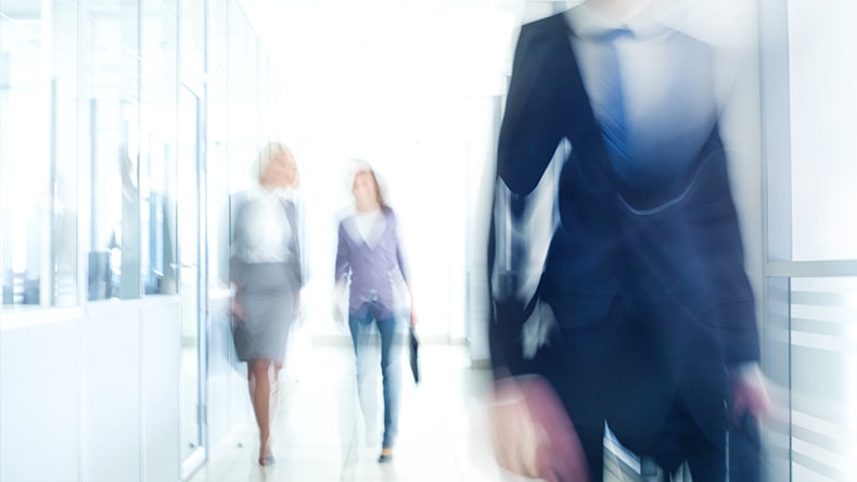 Blurred image of three people walking in a brightly lit office corridor. Two individuals in business attire walk towards the camera, while one person in a suit walks away, partially out of frame.